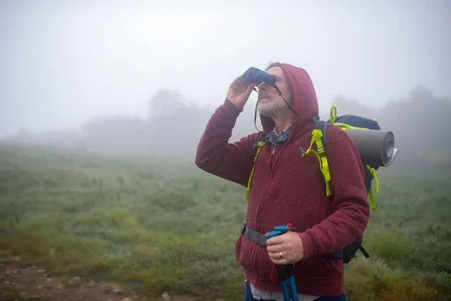 A bearded man with a backpack and trekking poles looking through binoculars on a foggy hike