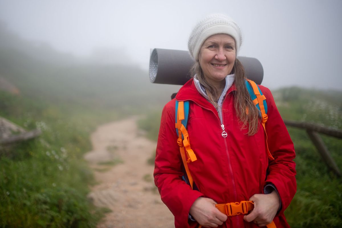 A smiling woman with a backpack and knit hat on a foggy hiking trail